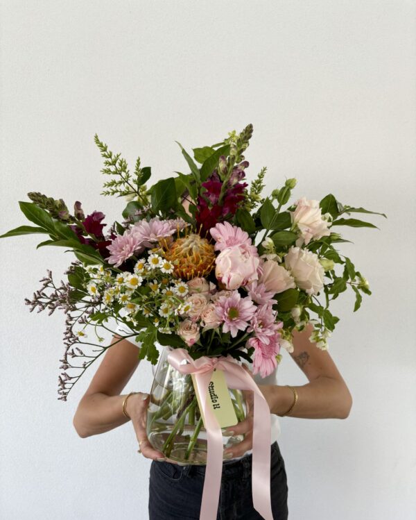 A person holds a large vase with a vibrant bouquet of pink gerberas and daisies.