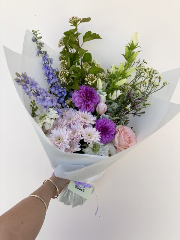A hand holds a 'Small Bunch' bouquet with purple dahlias, pink roses, and greenery.